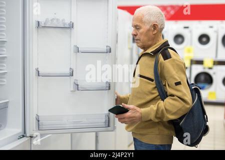 Älterer grauhaariger Rentner, der am Schalter im Ausstellungsraum der Abteilung für Elektrogeräte einen Kühlschrank sah Stockfoto