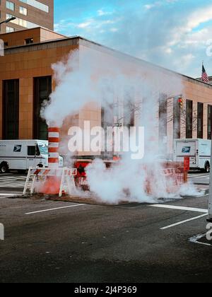 Rauchschwaden und Warnschild auf dampfender Straße bei einem Fußgängerüberweg mit uns Postautos im Hintergrund. Stockfoto