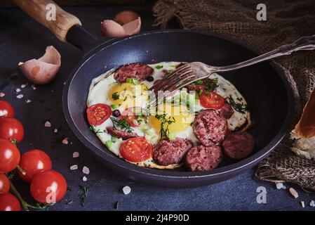 Spiegeleier mit geräucherter Wurst, Tomaten und Kräutern in einer Pfanne. Stockfoto