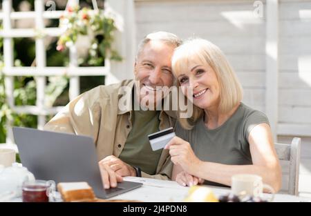 Online-Shopping-Konzept. Modernes Seniorenpaar mit Laptop und Kreditkarte, im Freien auf dem Land sitzend Stockfoto