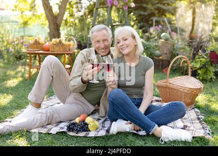 Alles Gute zum Jahrestag. Liebevolles verheiratetes Seniorenpaar, das Wein trinkt, während es auf der Decke sitzt und in seinem Garten ein Picknick macht Stockfoto