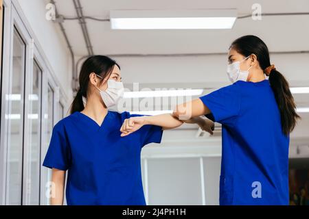 Junge weibliche Gesundheitshelferinnen, die eine OP-Maske tragen und die Hände mit Ellenbogenstoß schütteln, während sie während des Ausbruchs der Corona-Virus-Pandemie die soziale Distanz aufrechterhalten Stockfoto