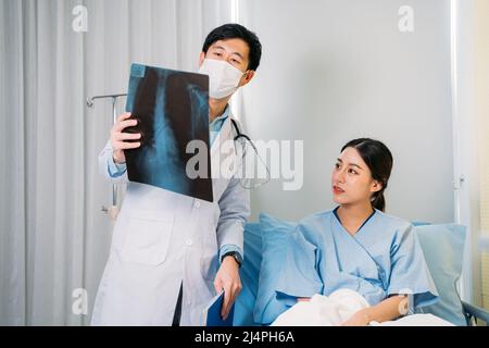 Junger asiatischer Arzt mit Gesichtsmaske und Stethoskop im Laborkittel, der den im Krankenhausbett sitzenden Patienten über den Röntgenbericht informiert und diesen überprüft Stockfoto
