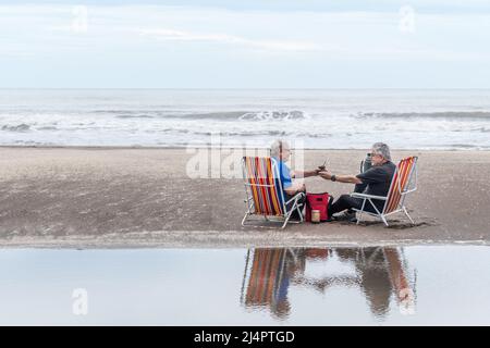 Zwei reife Männer mit grauen Haaren sitzen auf einem Strandstuhl trinken Mate alle im Wasser reflektiert. Hinter den Meereswellen. Stockfoto