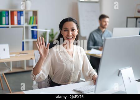 Glückliche junge Frau, die an einem modernen Computer arbeitet, lächelt und winkt der Kamera im Freiraum-Büro zu Stockfoto
