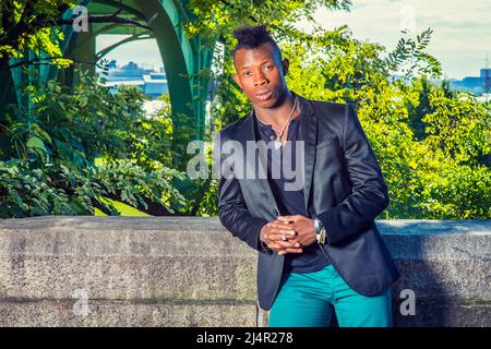 Mann, Der Sich Draußen Entspannt. In einem schwarzen Blazer, einem schwarzen Unterhemd, einer grünen Hose, steht ein junger schwarzer Kerl mit Mohawk-Haaren an einem felsigen Zaun, RE Stockfoto