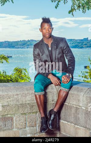 Mann, Der Sich Draußen Entspannt. In einem schwarzen Blazer, einer grünen Hose, Lederschuhen sitzt ein junger Schwarzer mit Mohawk-Haaren auf einem felsigen Zaun an einem riv Stockfoto