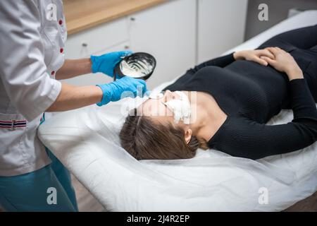 Arzt Kosmetologe oder Dermatologe macht Gesichtsmaske in Kosmetologie Salon. Professionelle Kosmetikerin, die Gesichtsmaske auf kaukasische Frau im Gesicht liegend Stockfoto