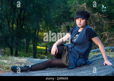 Ein junges chinesisches Mädchen sitzt ernsthaft in einer blauen, ärmellosen, langen Bluse, schwarzen Leggings und Stiefeln, in einem schwarzen Cord-Cop Stockfoto