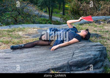 Ein junges chinesisches Mädchen liegt auf Felsen und liest in einer blauen, ärmellosen langen Bluse, schwarzen Leggings und Stiefeln, in einem schwarzen Cord-Cop Stockfoto