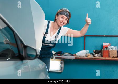 Das Porträt einer jungen lächelnden Mechanikerin in Gläsern und Overalls zeigt den Daumen hinter der Motorhaube eines Autos. Das Konzept der Frauenarbeit bei Männern Stockfoto