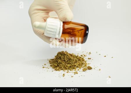 Marihuanaöl-Tropfflasche in den Händen eines Arztes mit zerdrücktem Marihuana im Stapel, weißer Hintergrund Stockfoto