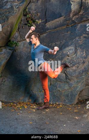 Ein junger Mann, der sich in einem grauen Langarm mit einem Henley-Hemd mit Rolllasche, roten Jeans und braunen Lederstiefelschuhen kleidet, streckt Arme und Beine, exercisi Stockfoto