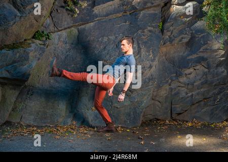 Ein junger Mann, der sich mit einem grauen Langarm mit einem Henley-Hemd mit Rolllasche, roten Jeans und braunen Lederstiefelschuhen kleidet, streckt die Beine und trainiert den Roboter Stockfoto