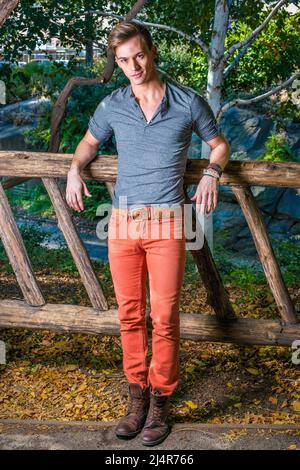Ein junger Mann, der ein graues Langarm mit einem Henley-Hemd mit Rolllasche, roten Jeans und braunen Lederstiefelschuhen trägt, steht charmant gegen einen Holzzaun Stockfoto