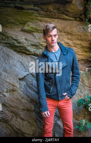 Ein junger Mann, der sich in einer blauen Slim-Fit-Jacke, einer roten Jeans ankleidet, steht bei Steinen, entspannt, denkt. Stockfoto