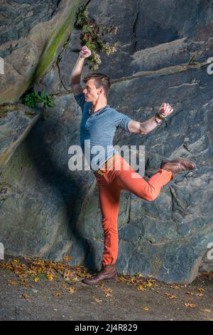 Ein junger Mann, der mit einem grauen Langarm, einem Henley-Hemd mit Rolllasche, roten Jeans und braunen Lederstiefelschuhen an den Armen und Beinen streckt und trainiert Stockfoto