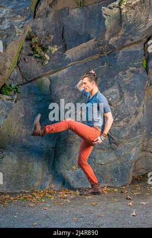 Ein junger Mann, der sich mit einem grauen Langarm mit einem Henley-Hemd mit Rolllasche, roten Jeans und braunen Lederstiefelschuhen kleidet, dehnt die Beine, wahrscheinlich ein wandelbarer Roboter Stockfoto