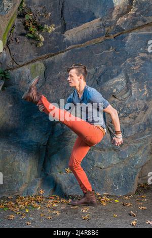 Ein junger Mann, der sich mit einem grauen Langarm mit einem Henley-Hemd mit Rolllasche, roten Jeans und braunen Lederstiefelschuhen kleidet, dehnt die Beine, wahrscheinlich ein wandelbarer Roboter Stockfoto