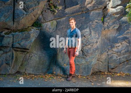 Ein junger Mann, der ein graues Langarm mit einem Henley-Hemd mit Rolllasche, roten Jeans und braunen Lederstiefelschuhen trägt, steht an den großen Felsen, lächelt und sieht aus Stockfoto