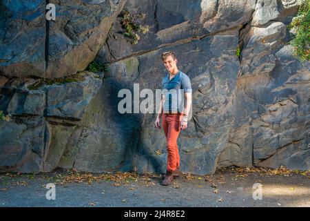 Ein junger Mann, der ein graues Langarm mit einem Henley-Hemd mit Rolllasche, roten Jeans und braunen Lederstiefelschuhen trägt, steht an den großen Felsen, lächelt und sieht aus Stockfoto