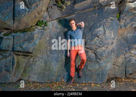 Ein junger Mann lehnt sich an große Felsen und schaut dich an, trägt graue lange Ärmel mit Henley-Hemd mit Rolllasche, roten Jeans und braunen Lederstiefelschuhen. Stockfoto