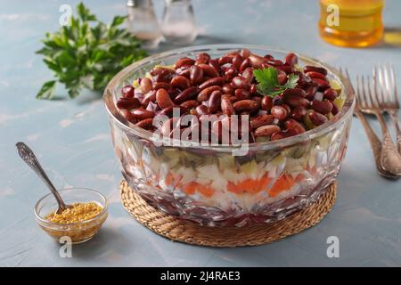 Geschichtete Vinaigrette mit roten Bohnen, Gurken, gekochten Kartoffeln, Rüben und Karotten in einer transparenten Salatschüssel auf blauem Hintergrund Stockfoto