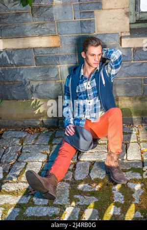 Mann Casual Fashion. In blauem, weißem Muster-Hemd, einer Kapuzenjacke, roten Jeans, Lederstiefelschuhen, Hält einen wollenen Fedora-Hut auf dem Knie, ein Jo Stockfoto
