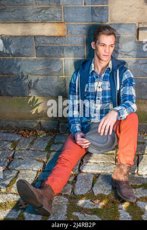 Mann Casual Fashion. In blauem, weißem Muster-Hemd, einer Kapuzenjacke, roten Jeans, Lederstiefelschuhen, Mit einem wollenen Fedora-Hut ist ein junger Mann s Stockfoto