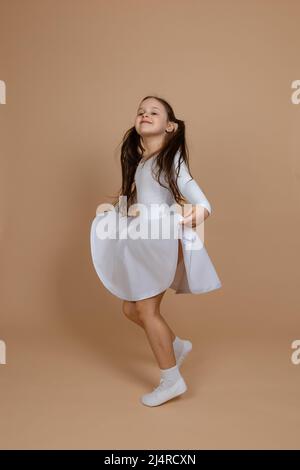 Seitenansicht eines jungen lächelnden Mädchens mit langen dunklen Haaren in weißem Kleid, Socken und Turnschuhen stehend, Kleid mit Händen haltend, Spaß auf Braun Stockfoto