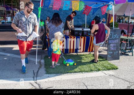 Blondes Kind mäht mit einem Spielzeug-Rasenmäher vor einem Taco-Stand einen Fleck künstliches Gras, während Erwachsene bei Cherry Street Farme Lebensmittel kaufen Stockfoto