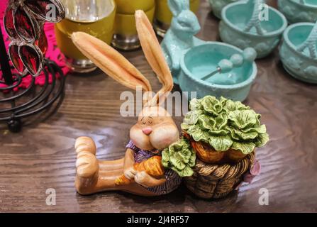 Osterdekor und -Geschirr - schlafender Keramikkaninchen mit verschwommenen Ohren hält Karotten auf der Holzoberfläche fest, die von Hasenschüsseln umgeben ist Stockfoto