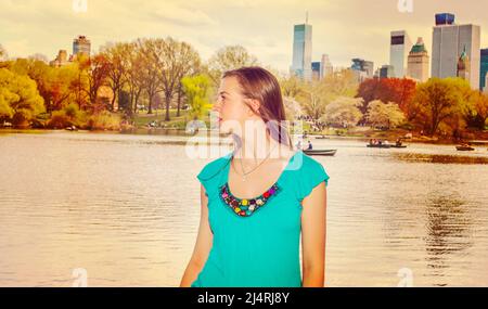 Das amerikanische Teenager-Mädchen, das in blauer ärmelloser Kleidung in New York unterwegs ist, steht am Frühlingstag am See im Central Park und schaut sich um. Personen r Stockfoto