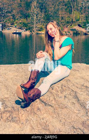 American Teenage Girl trägt ein blaues ärmelloses Oberteil, modische Jeans, braune Stiefel, sitzt auf Felsen am See im Central Park, New York, und spricht über Zelle Stockfoto