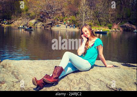 American Teenage Girl trägt ein blaues ärmelloses Oberteil, modische Jeans, braune Stiefel, sitzt auf Felsen am See im Central Park, New York, und spricht über Zelle Stockfoto