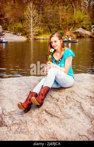 Das amerikanische Teenager-Mädchen trägt ein blaues ärmelloses Oberteil, Jeans, braune Stiefel, sitzt auf Felsen am See im Central Park, New York, hält weiße Blume, riecht Stockfoto