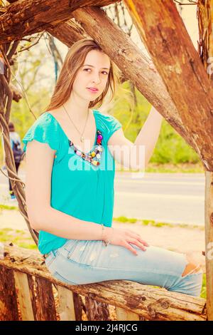 Das amerikanische Teenager-Mädchen trägt ein blaues ärmelloses Oberteil, modische Jeans und sitzt auf einem Holzzaun im Central Park, New York, und blickt auf Sie charmant. Zoll Stockfoto