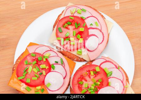 Knusprige Cracker Sandwiches mit Tomaten, Wurst, Käse, grünen Zwiebeln und Radish auf weißem Teller. Einfaches Frühstück. Schnelle und gesunde Sandwiches. Knäckebrot mit leckerer Füllung. Gesunder Ernährungs-Snack Stockfoto