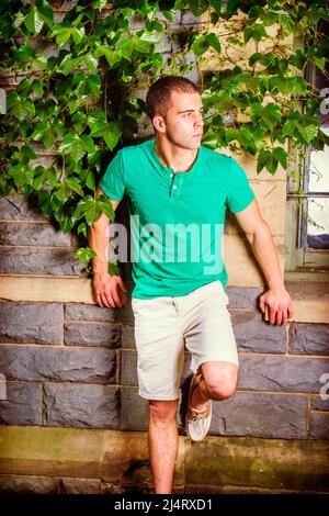 Junger Mann wartet. Ein junger Mann, der ein grünes, kurzärmeliges Henley-Hemd trägt, hellgelbe Shorts trägt und ein Bein beugt, steht mit Grün gegen die Wand Stockfoto