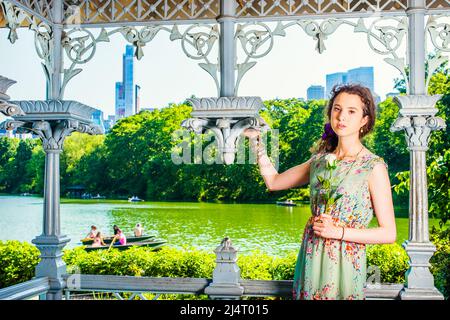 Junge Dame wartet auf Euch. Hübsches Mädchen im Teenageralter in ärmellosem, langem Kleid, im Pavillon im Park stehend, mit der Hand die weiße Rose haltend und anblickend Stockfoto