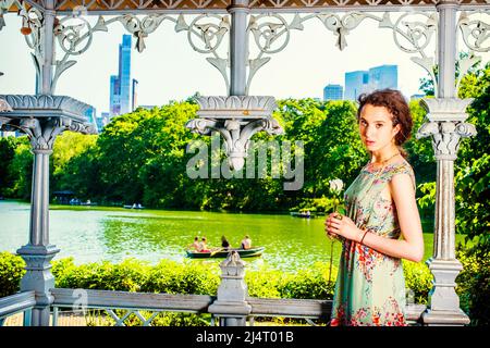 Junge Dame wartet auf Euch. Hübsches Mädchen im Teenageralter in ärmellosem, langem Kleid, im Pavillon im Park stehend, mit der Hand die weiße Rose haltend und anblickend Stockfoto