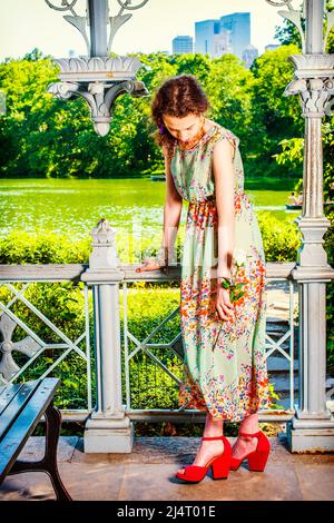 Sommermode. In ärmellosen, langen Kleidern, roten Sandalen-Schuhen kleidet, steht ein Teenager-Mädchen in einem Garten, eine Hand mit einer weißen Rose, loweri Stockfoto