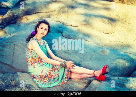 Sommermode. Schöne Teenager-Mädchen trägt ärmelloses, langes Kleid, rote Sandalen Schuhe, lila Blume Haarnadel, klobige Kette Armband, Sitzen auf BI Stockfoto