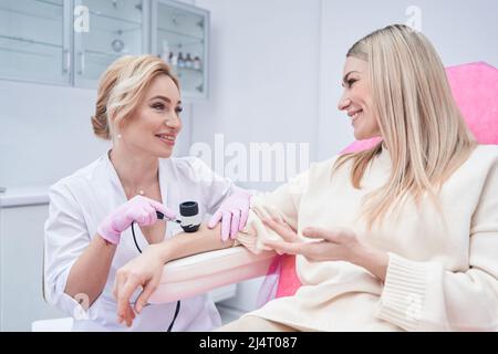 Fröhlicher Patient, der während der Dermatoskopie mit dem Dermatologen kommuniziert Stockfoto