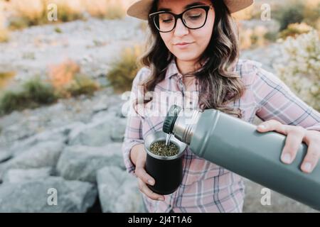 Junge latinerin mit Hut, serviert Mate mit einem hellgrünen Thermo, in einem natürlichen Raum. Stockfoto