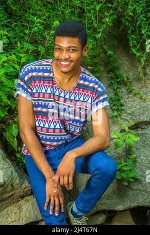 Porträt des jungen schwarzen Mannes. Ein junger, gutaussehender Kerl sitzt mit einem kurzen, kragenlosen, farbenfrohen Musterglüt gegen Felsen mit grünem Lauch Stockfoto