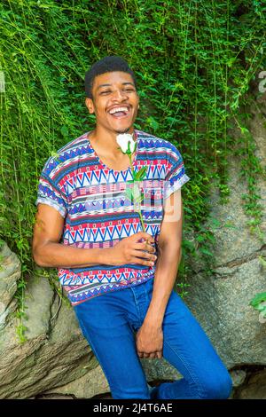 Glücklicher Mann. In einem kurzärmeligen, kragenlosen, farbenfrohen Musterkhemd, einer blauen Jeans sitzt ein junger Schwarzer gegen Felsen mit grünen Blättern, Holdi Stockfoto