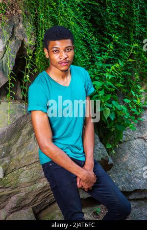 Porträt des jungen schwarzen Mannes. Tragen eines grünen, kurzen Ärmeln, V-Ausschnitt T-Shirt, schwarze Hose, Hände halten, Ein junger, gutaussehender Kerl lehnt sich an Felsen Stockfoto