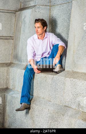 Mann, Der Draußen Arbeitet. Ein junger Geschäftsmann sitzt in einem hellrosa, langärmeligen Hemd, blauen Jeans und Lederschuhen lässig gegen eine Betonwa Stockfoto