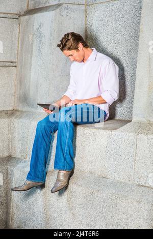 Mann, Der Draußen Arbeitet. Ein junger Geschäftsmann sitzt in einem hellrosa, langärmeligen Hemd, blauen Jeans und Lederschuhen lässig gegen eine Betonwa Stockfoto